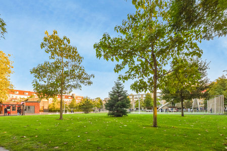 a park with trees and people walking on the sidewalk in the distance is a red brick building that has been builtのeditorial素材