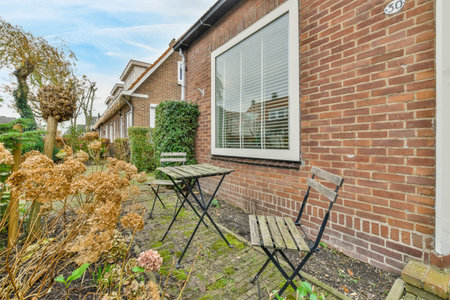 the outside of a house with some plants and flowers in front of it, there is an empty chair by the...