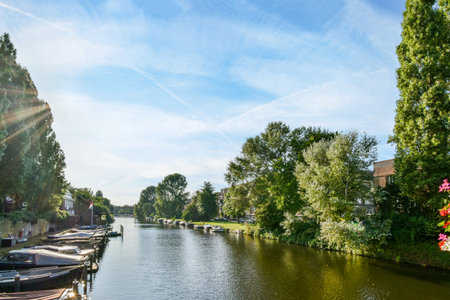 Amsterdam, Netherlands - 10 April, 2021: some boats in the water near trees and buildings with sun shining through the blue sky over them on a sunny dayのeditorial素材