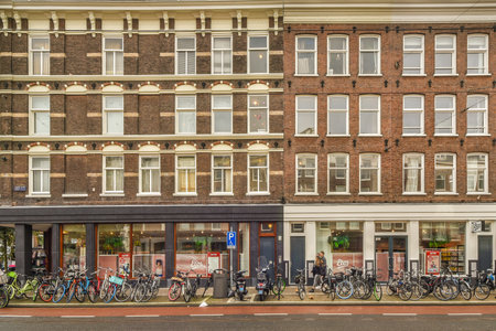 Amsterdam, Netherlands - 10 April, 2021: some bikes parked in front of a large brick building with many windows on the side of the street and people walking down the sidewalkのeditorial素材