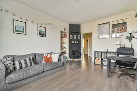 a living room with wood flooring and gray couch in the center of the room is an office desk,...