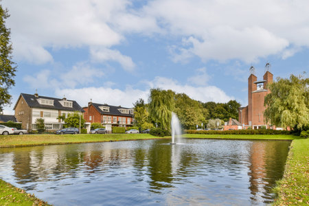 A tranquil park scene featuring a serene pond with a fountain, surrounded by lush greenery and charming buildings under a blue sky. Ideal for relaxation.の写真素材