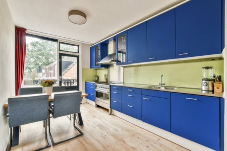 A contemporary kitchen featuring sleek blue cabinets, a tidy dining area, and large windows that offer natural light and views of greenery outside.の写真素材