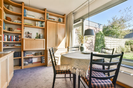 An inviting indoor living area featuring a stylish wooden bookshelf, a dining table with chairs, and large windows allowing natural light to illuminate the space.の写真素材