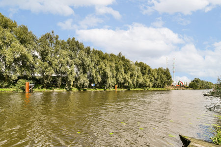 A tranquil river scene featuring greenery lining the banks, with distant structures and a clear blue sky enhancing the peaceful atmosphere.の写真素材