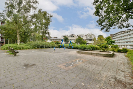 An empty playground area featuring swings, benches, and patches of grass, surrounded by residential buildings under a cloudy sky.の写真素材