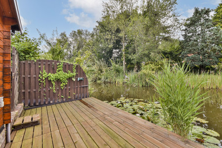 A serene garden scene featuring lush greenery surrounding a tranquil pond, wooden deck, and a rustic wooden gate, inviting a peaceful atmosphere.の写真素材