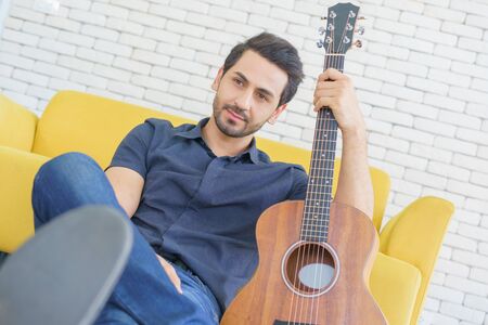 Happy man playing guitar while sitting on sofa in living room, Enjoying carefree time at home.