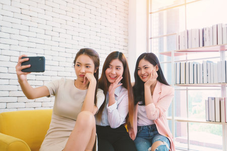 Beautiful young business women are selfie with her friend by cellphone or smartphone at living room. Happy friendship relationship concept.の写真素材
