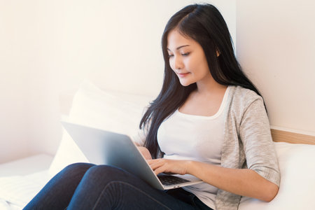 Young woman working on a laptop sitting on bed at home.の写真素材