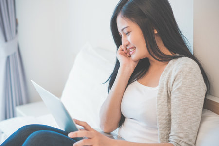Young woman working on a laptop sitting on bed at home.の写真素材