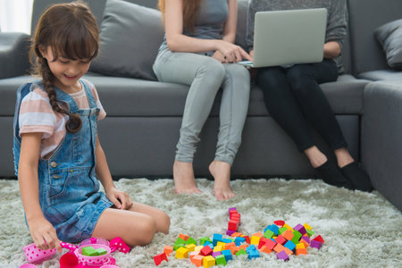 child a little girl is playing blocks at home. Mother with friend working on laptop or shopping online.の写真素材