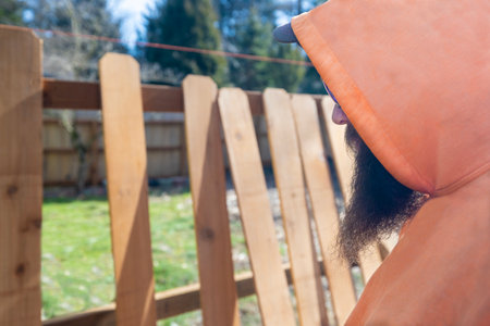 Fence Builder Construction Worker With Beard Examines and Measures Wood Pickets Along a Grass Yardの写真素材