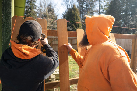 Two Construction Workers Build an Outdoor Fence One Man Helping Another Line Up Wood Picket Slats on Level Angle Secured By Screws With Power Toolの写真素材