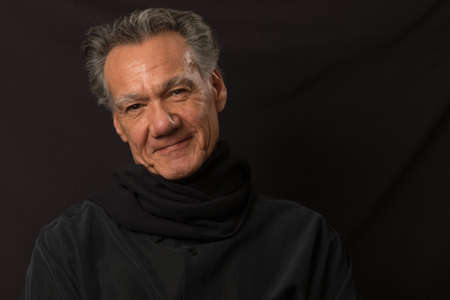 Portrait of an older man smiling wearing a black shirt and scarf around his neck against a black...