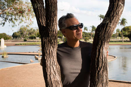 Senior man wearing dark shades at the park standing behind a tree on a sunny dayの写真素材