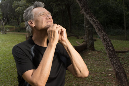 Mature Man outside at a park sitting on a tree stump stretching out his neck for pain relief