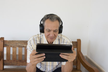 Mature Man outside sitting on a bench watching a show on his iPad Smart Tablet wearing headphonesの写真素材