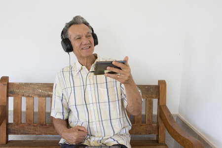 Mature Man outside sitting on a bench watching a show on his Smart Phone wearing headphonesの写真素材