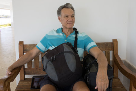 Mature Man sitting on a bench with his suitcase and backpack waiting for a rideの写真素材