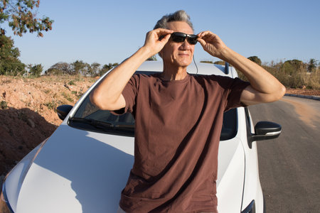 Mature Man taking a break from driving and relaxing and leaning on the hood of his car