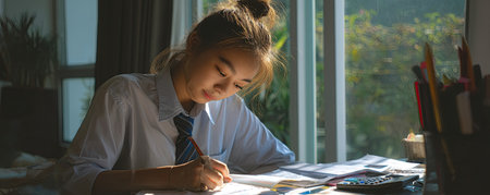 A young woman in a school uniform writes at her desk, bathed in sunlight near a window, studying.の素材