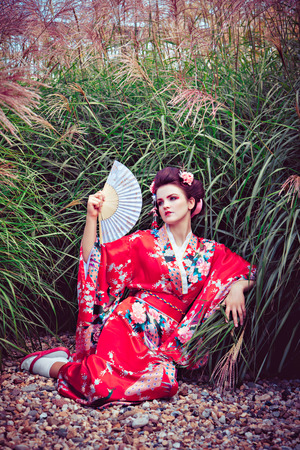 Young sitting woman in geisha costume with fancy makeup in garden with a fanの写真素材