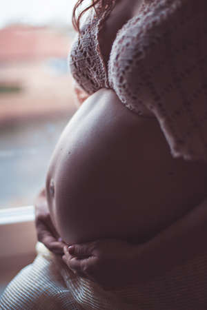Close-up of pregnant woman with hands under tummy at window backgroundの写真素材
