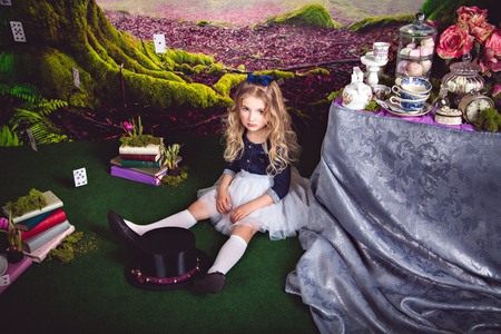 Little sitting on the floor girl in a beautiful dress in the image of Alice in Wonderland and magician hat in front of her