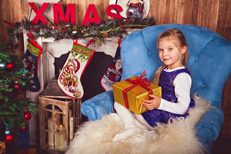 Girl with gift in festive beautiful clothes celebrating Christmas in decorated room on the background of the Christmas tree and fireplace. Studio photoの写真素材