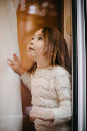 Lonely little girl waiting near the window inside the room during quarantine and lockdown in a pandemicの写真素材