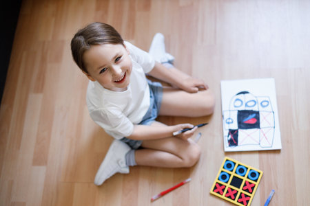 Happy little child is sitting comfortably on the wooden floor, drawing on paper with colored pencilsの写真素材