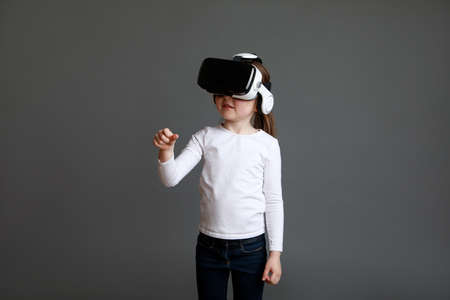 Fascinated female child in white long sleeve shirt looking into virtual reality glasses over gray background.の写真素材