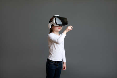 Little preschool girl experiencing virtual reality eyeglassses in white long sleeve shirt over gray background.の写真素材