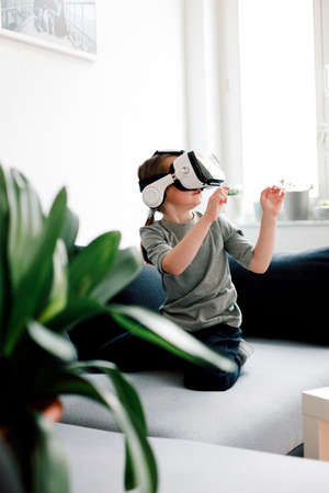 Funny cute girl watching something that is displayed in goggles of virtual reality, sitting on the couch. Modern technology conceptの写真素材