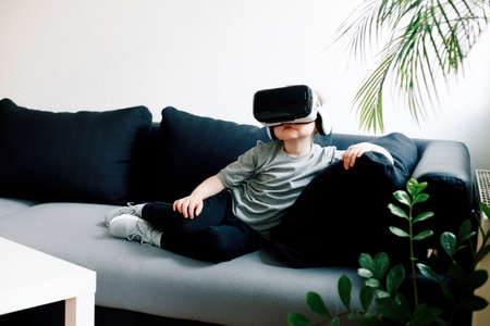 Funny cute girl watching something that is displayed in goggles of virtual reality, lying on the couch. Modern technology conceptの写真素材