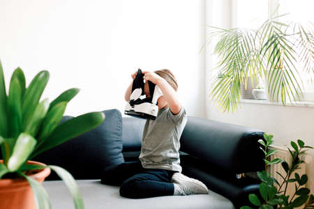 Funny cute girl watching something that is displayed in goggles of virtual reality, sitting on the couch. Modern technology conceptの写真素材