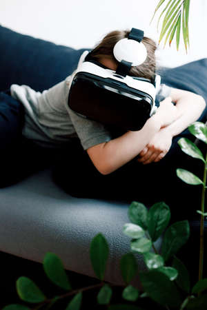 Funny cute girl watching something that is displayed in goggles of virtual reality, lying on the couch and holding pillow. Modern technology conceptの写真素材