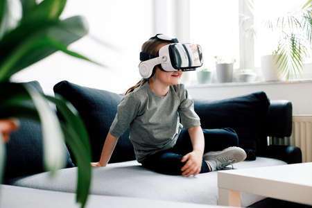 Funny cute girl watching something that is displayed in goggles of virtual reality, sitting on the couch. Modern technology conceptの写真素材