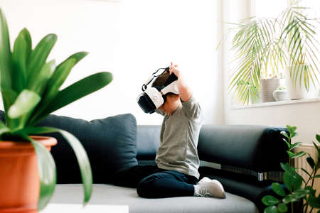 Funny cute girl watching something that is displayed in goggles of virtual reality, sitting on the couch. Modern technology conceptの写真素材
