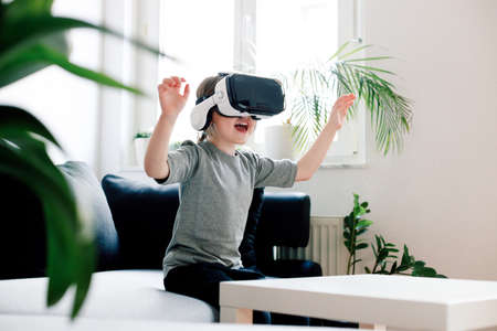 Funny cute girl watching something that is displayed in goggles of virtual reality, sitting on the couch. Modern technology conceptの写真素材