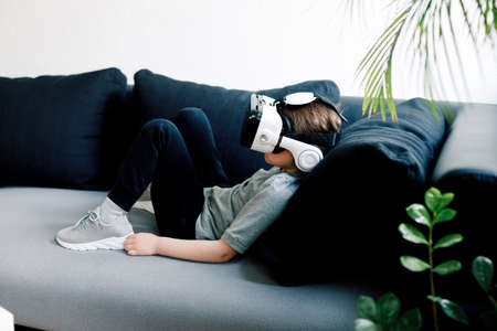 Funny cute girl watching something that is displayed in goggles of virtual reality, lying on the couch. Modern technology conceptの写真素材
