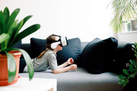 Funny cute girl watching something that is displayed in goggles of virtual reality, lying on the couch. Modern technology conceptの写真素材