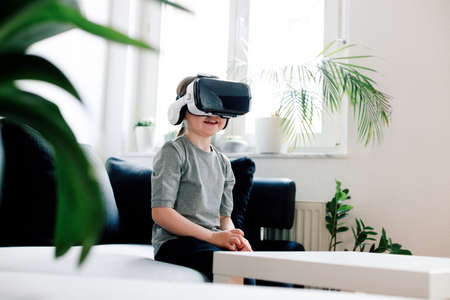 Funny cute girl watching something that is displayed in goggles of virtual reality, sitting on the couch. Modern technology conceptの写真素材