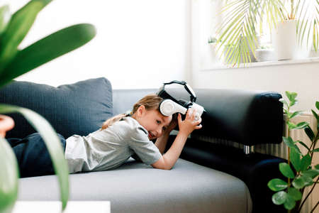 Funny cute girl watching something that is displayed in goggles of virtual reality, lying on the couch. Modern technology conceptの写真素材