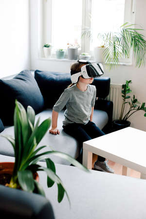 Funny cute girl watching something that is displayed in goggles of virtual reality, sitting on the couch. Modern technology conceptの写真素材