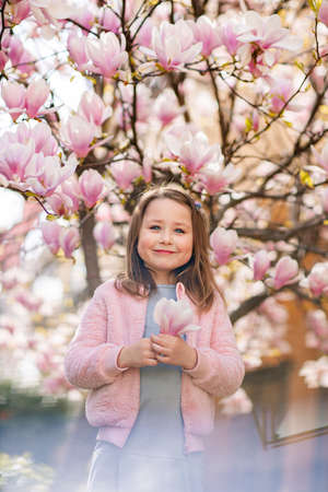 Portrait of a cute cheerful girl in a gray dress wearing rosa jacket staying in spring in a park under a flowering pink magnolia tree.の写真素材