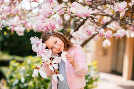 Portrait of a cute cheerful girl in a gray dress wearing rosa jacket staying in spring in a park under a flowering pink magnolia tree.の写真素材