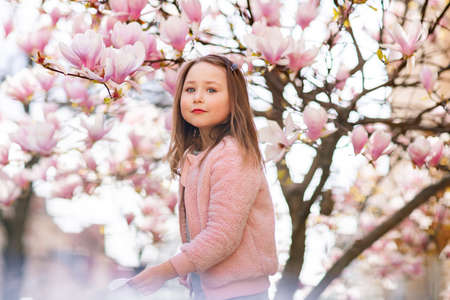 Portrait of a cute cheerful girl in a gray dress wearing rosa jacket staying in spring in a park under a flowering pink magnolia tree.の写真素材