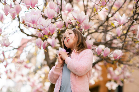 Portrait of a cute cheerful girl in a gray dress wearing rosa jacket staying in spring in a park under a flowering pink magnolia tree.の写真素材